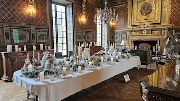 Een lange eettafel gedekt met kaarsen en wit servies, omringd door sierlijk meubilair en een open haard.