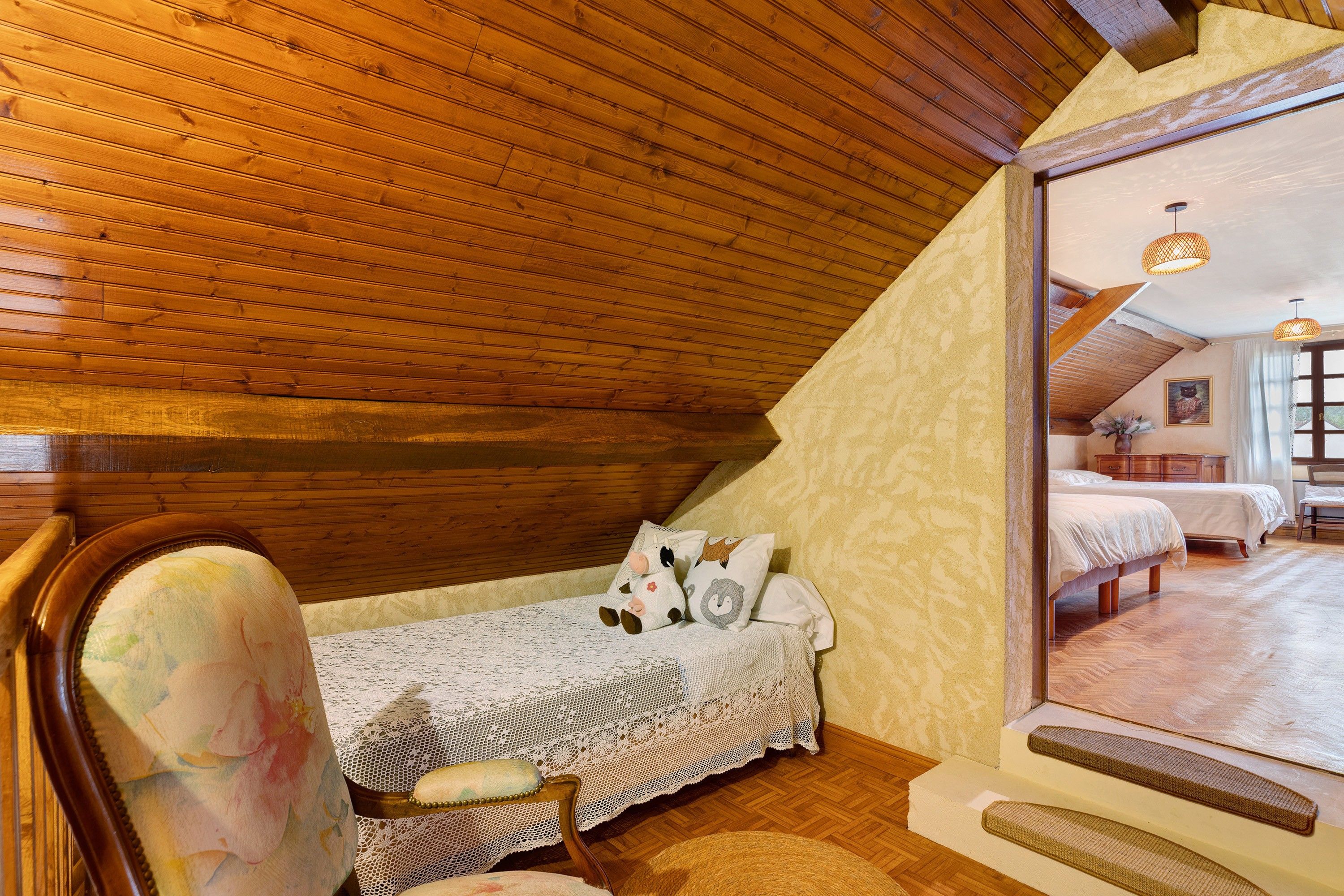 A cozy attic bedroom with a wooden sloped ceiling, a small bed with a white bedspread, and a chair with a floral cushion.
