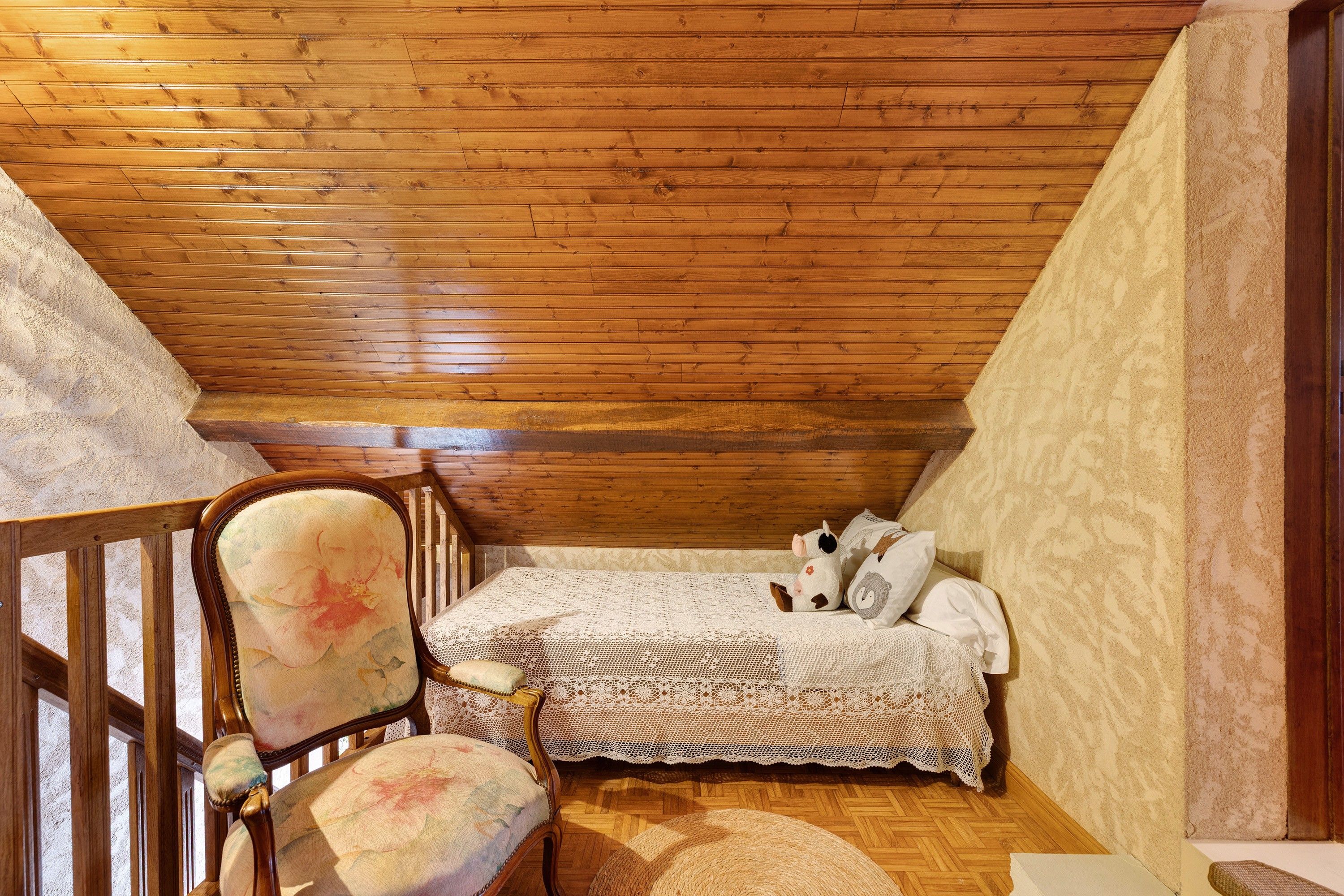 A cozy attic bedroom with wooden beams, a bed with a lace cover, a chair with floral upholstery, and a small rug.