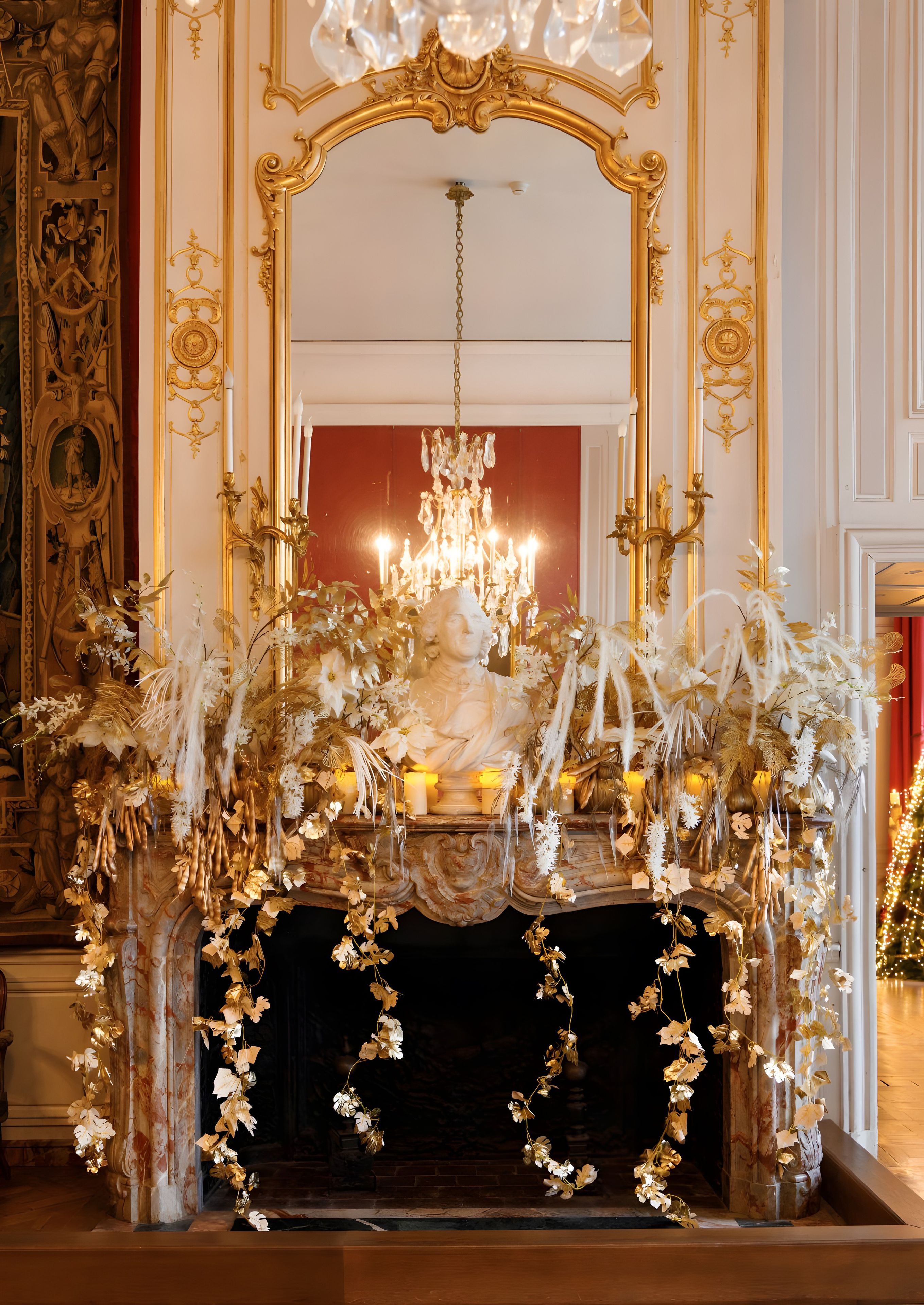 An ornate fireplace with a chandelier, surrounded by elaborate gold decorations and white floral arrangements.