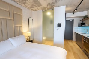 Una habitación de hotel con cama, lámpara de noche, espejo, teléfono y kitchenette.