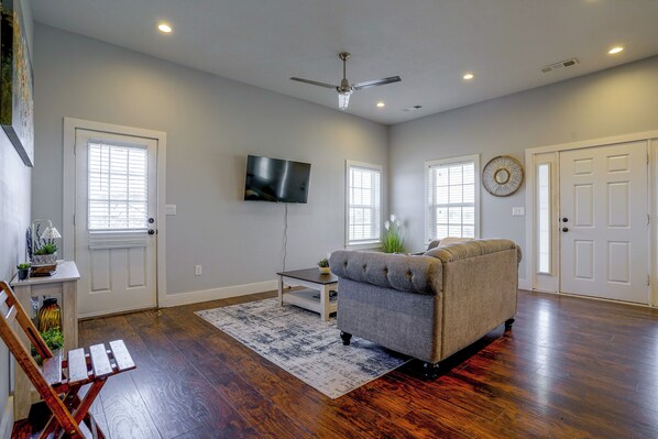 A living room with hardwood flooring, a grey sofa, a flat-screen TV mounted on the wall, and a ceiling fan. - Countryside Home Right Outside Fort Worth (Joshua)