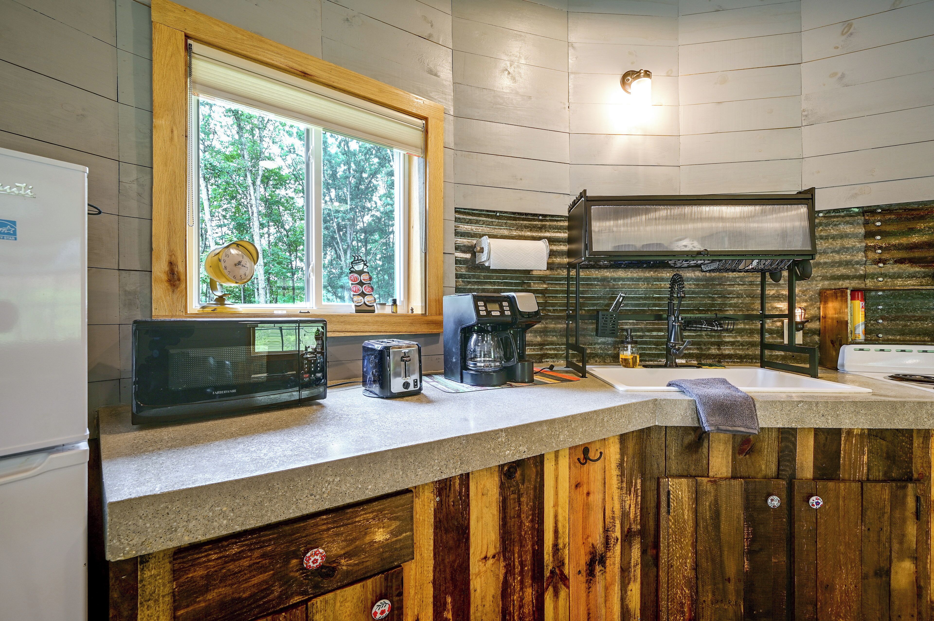 A kitchen with a refrigerator, microwave, toaster, coffee maker, and a window with a view of trees.