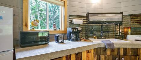 A kitchen with a refrigerator, microwave, toaster, coffee maker, and a window with a view of trees.