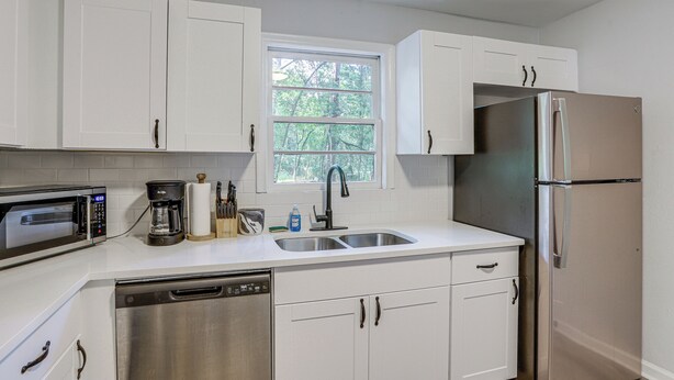 A kitchen with white cabinets, a stainless steel refrigerator, a microwave, a coffee maker, and a window with a view of trees.