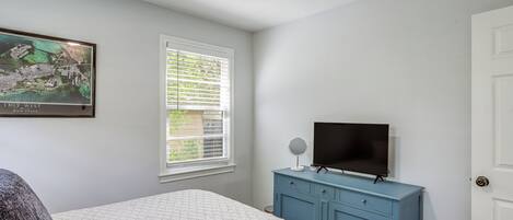 A bedroom with a bed, a dresser, a television, a ceiling fan, a window with blinds, and a framed picture on the wall.