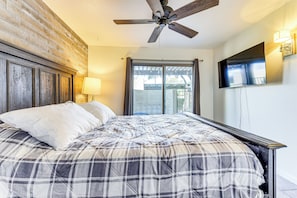 Ein Schlafzimmer mit einem Bett, einem Deckenventilator, einem Fenster mit Vorhängen und einem an der Wand befestigten Flachbildfernseher.