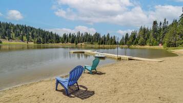 A lakeside view with a sandy beach, two chairs, and a dock extending into the water.