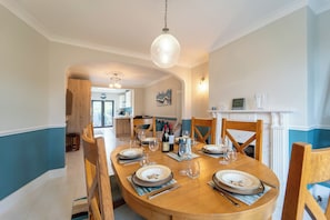 A dining room with a round table set for six, a fireplace, and a kitchen in the background. - Extended Family Home with Parking near Heathrow (Ickenham)