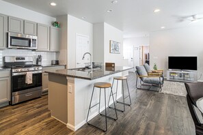 A modern kitchen with built-in appliances, a central island with stools, a dining area with a sofa and TV, and a hallway leading to other rooms. - 2BR, 2BA, 2Car - Office, Laundry, Quiet, by DIA (Denver)