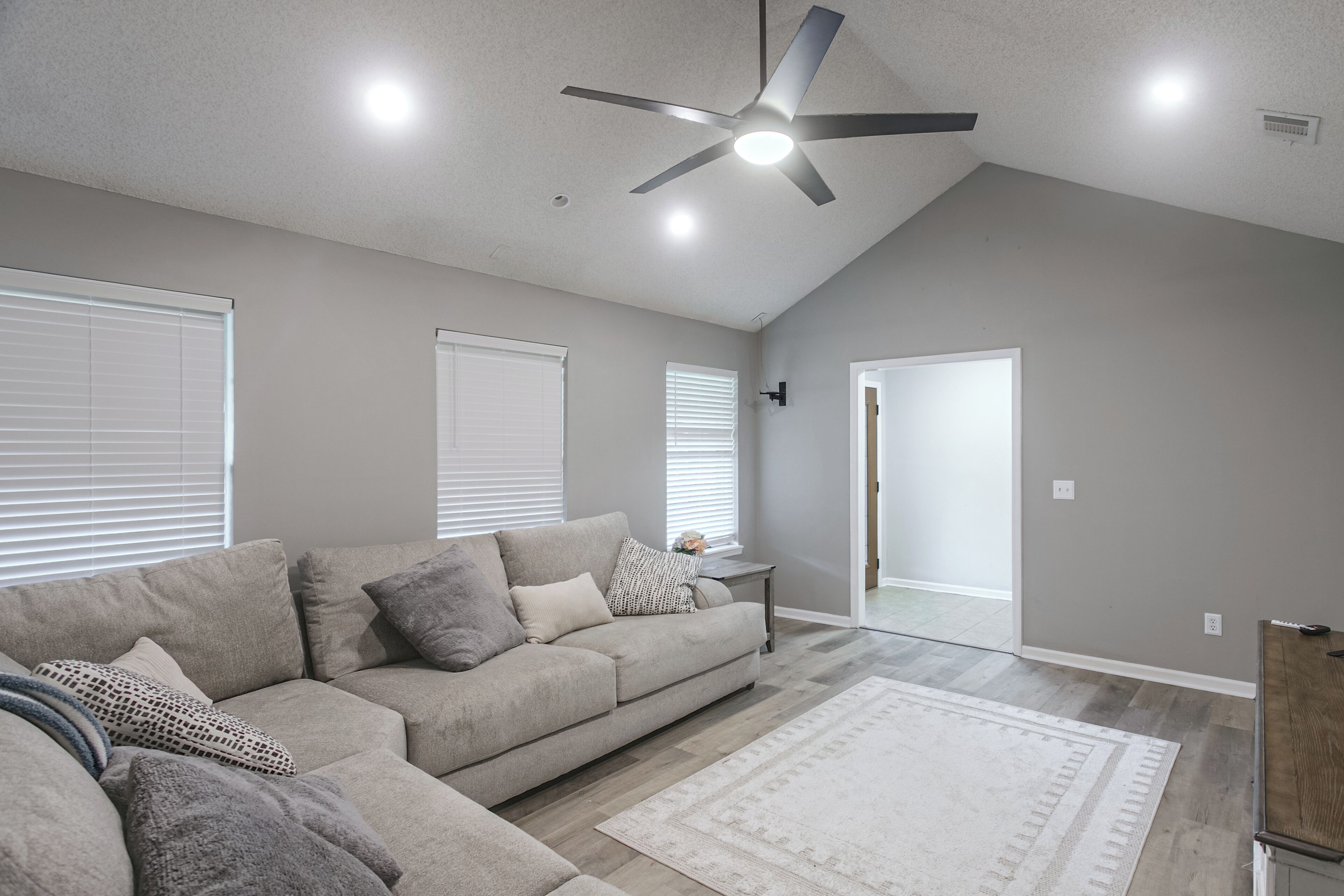 A modern living room with a sectional sofa, a ceiling fan, and a large window with blinds.