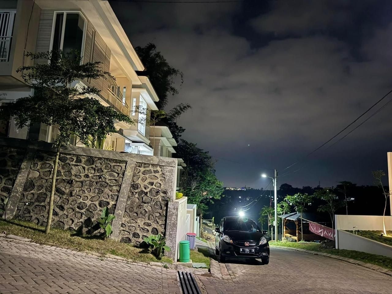 A residential street at night with a parked car, a stone wall, and a building with a balcony.