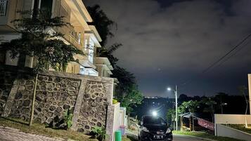 A residential street at night with a parked car, a stone wall, and a building with a balcony.