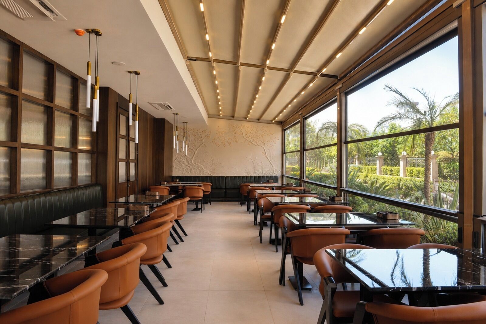 Un restaurant amb una barra llarga, diverses taules i cadires. Grans finestres ofereixen vistes a zones verdes a l'exterior.