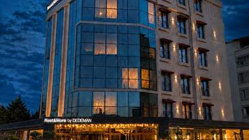 A multi-story hotel with a prominent 'Rest&More by DEDEMAN' sign, featuring a modern architectural design with large windows and a well-lit entrance.