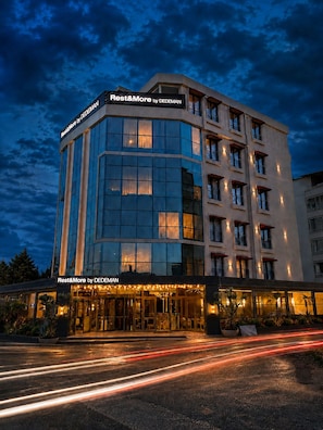 A multi-story hotel with a prominent 'Rest&More by DEDEMAN' sign, featuring a modern architectural design with large windows and a well-lit entrance.
