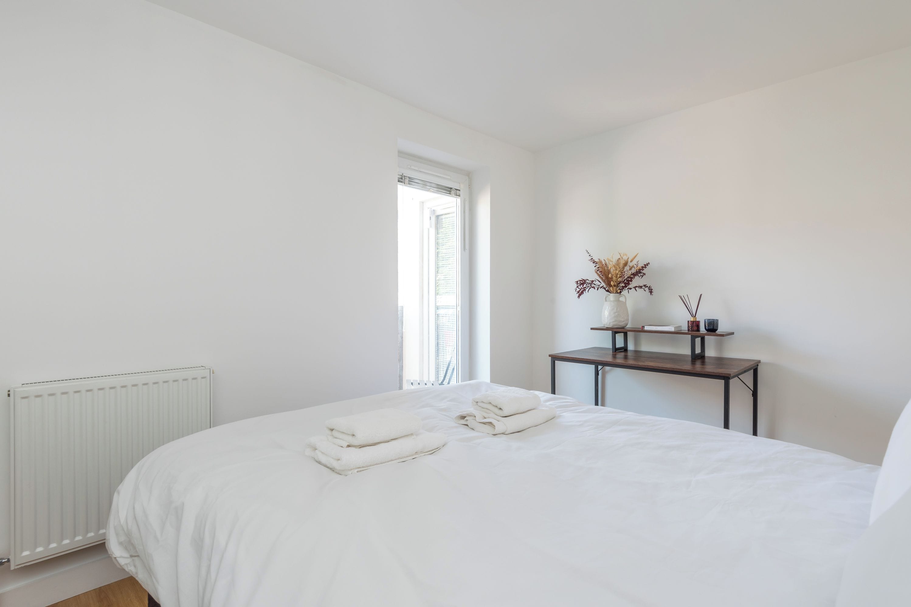 Een minimalistische slaapkamer met een bed, een radiator, een klein tafeltje met decoratieve objecten en een raam.