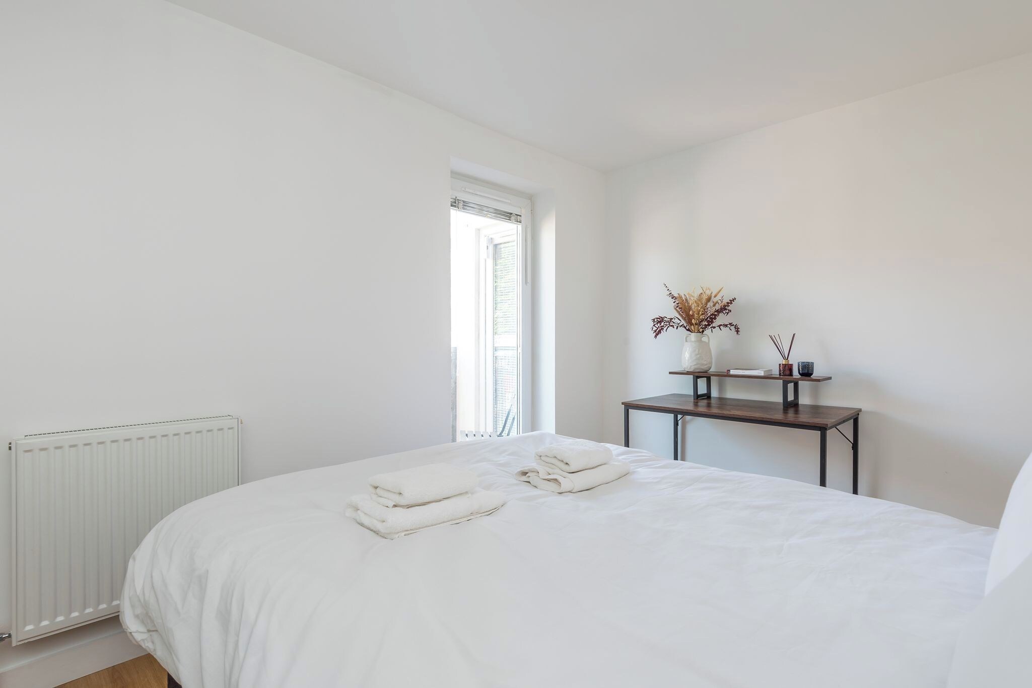 A bedroom with a bed, a bedside table with a vase and a book, and a window with blinds.