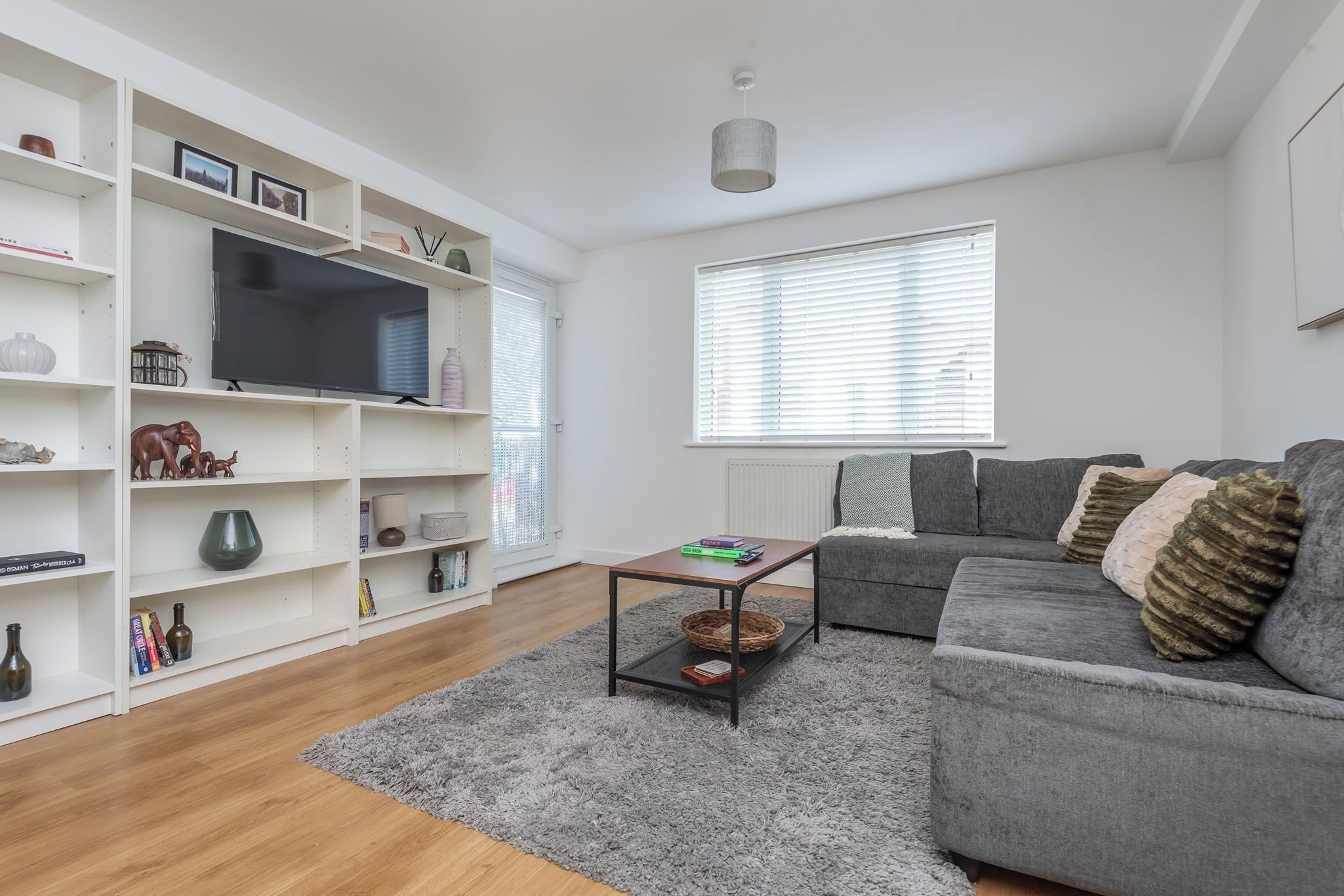 A modern living room with a grey sofa, a flat-screen TV on a white shelf, and a grey rug.