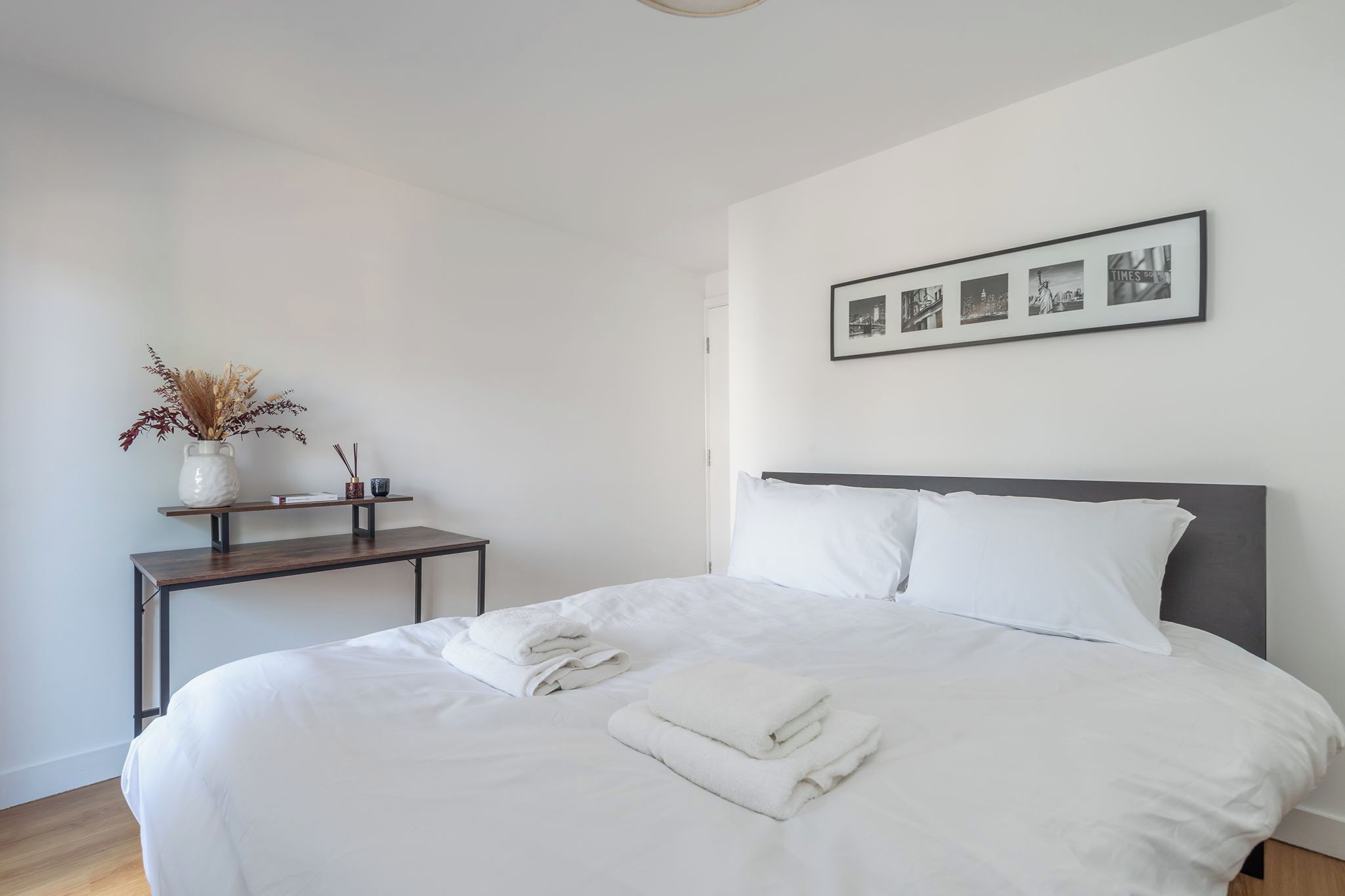 A neatly made bed with white linens and folded towels, a small bedside table with a vase and a cup, and a framed picture of cityscapes on the wall.