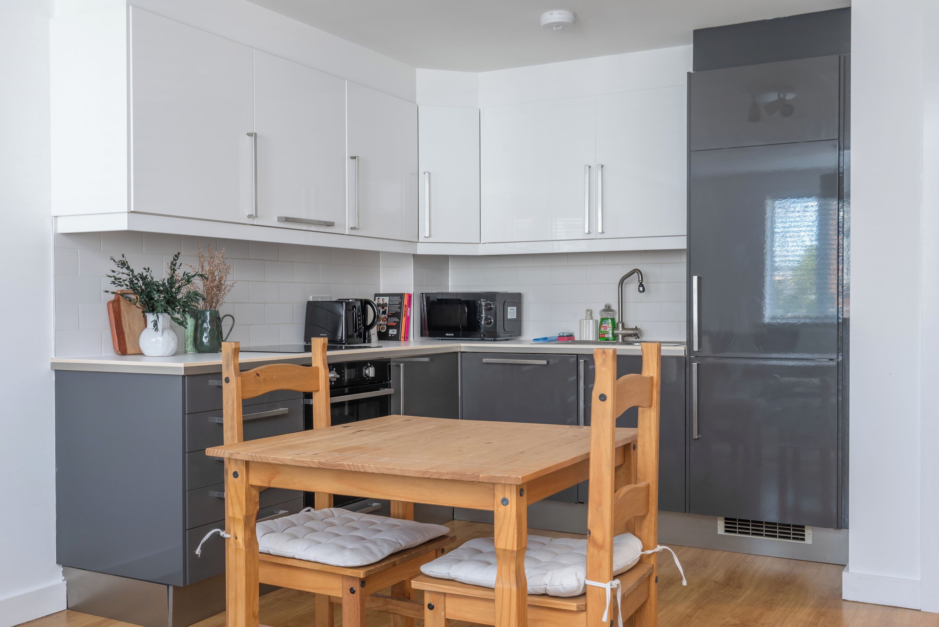 A modern kitchen with white cabinets, a black refrigerator, a microwave, and a wooden table with cushioned chairs.