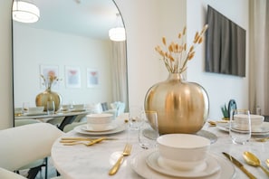 Ein Esszimmertisch ist mit weißen Tellern, Schüsseln und goldbestecktem Besteck gedeckt, in der Mitte steht eine große Vase mit getrockneten Pflanzen.