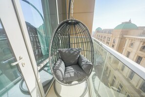 En hængestol med hynder på en balkon med gelænder af glas, med udsigt over en by med klassisk arkitektur.