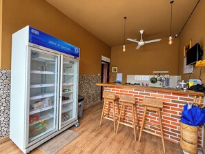 A kitchen area with a GEA refrigerator, a bar counter with stools, a ceiling fan, and a wall-mounted TV. - Pondok Juli Canggu (Canggu)