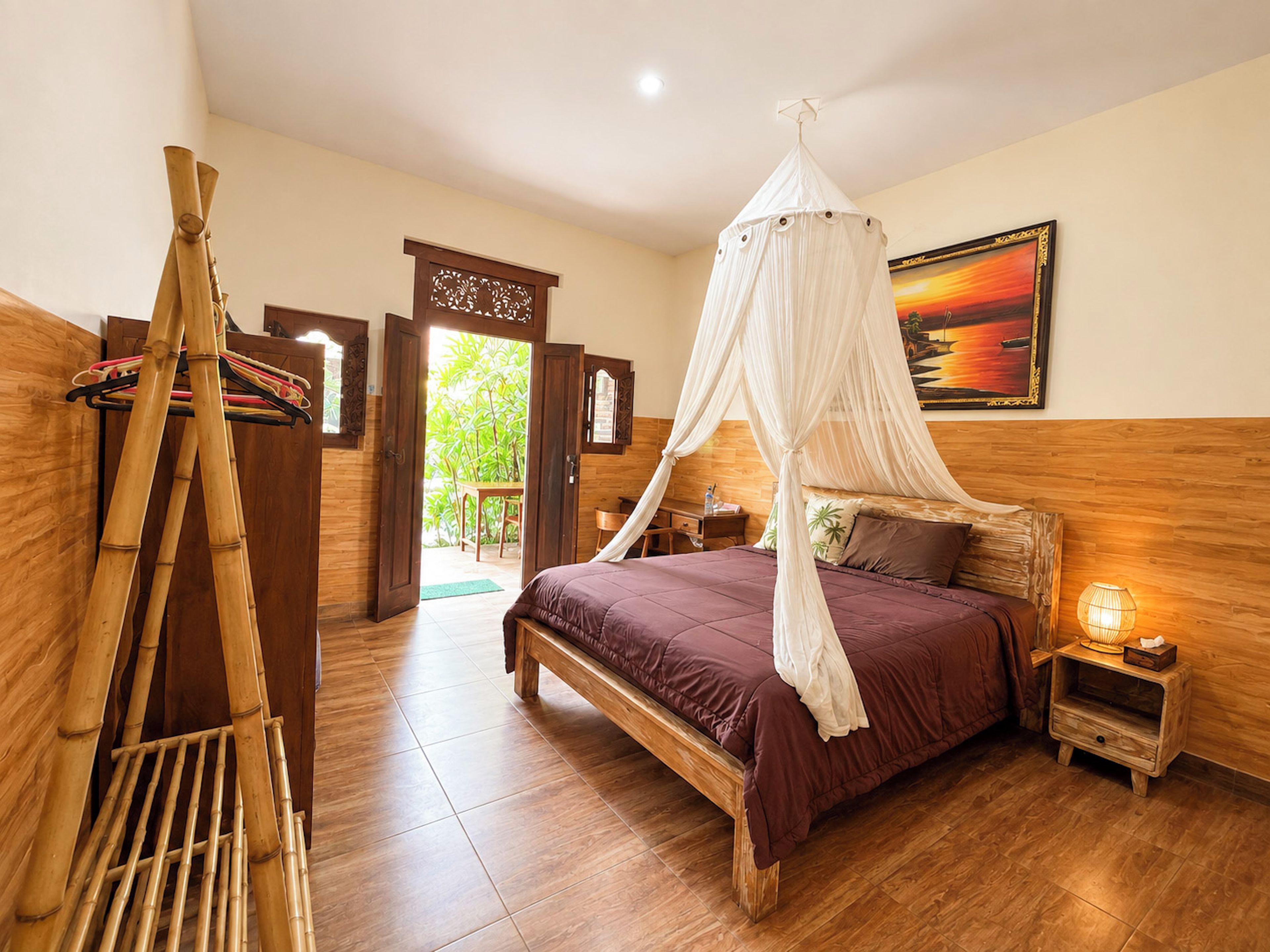 A room with a bed, a nightstand, a wooden floor, a door leading outside, and a wall with a framed picture.