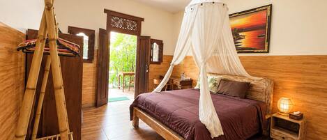 A room with a bed, a nightstand, a wooden floor, a door leading outside, and a wall with a framed picture.
