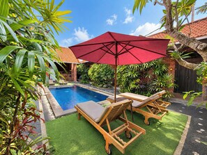 Un espace piscine agrémenté d’un parasol rouge, de chaises longues en bois et d’une végétation luxuriante.