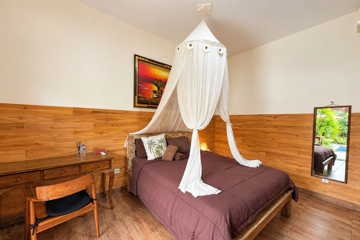 Een slaapkamer met een hemelbed, een houten hoofdbord, een bureau met stoel en een schilderij aan de muur.