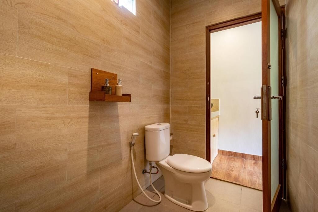 Un baño con inodoro, un estante de madera con artículos de aseo, y una puerta que da a otra habitación.