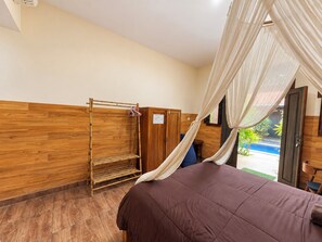 A room with a bed, a wooden wardrobe, a shelf unit, and a window with a view of a pool. - Pondok Juli Canggu (Canggu)