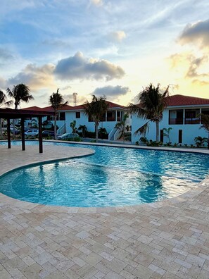 A swimming pool with a curved edge, surrounded by a paved walkway, palm trees, and buildings with red-tiled roofs. - New! Luxury 2-bedroom apartment with shared pool, only 5 minutes to Mambo Beach (Unknown)