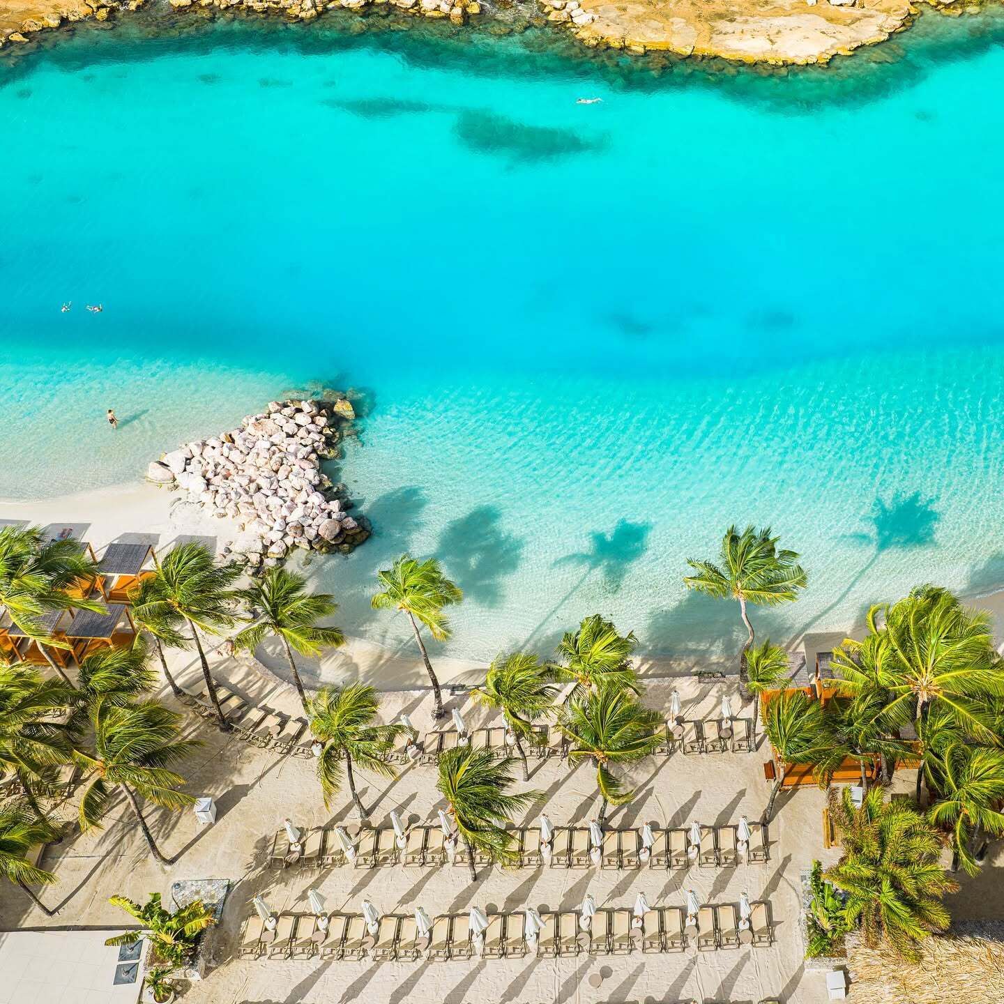 A beach resort with clear turquoise waters, a rocky breakwater, and rows of sun loungers and palm trees.