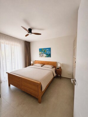 A hotel room with a wooden bed, a ceiling fan, a painting of a beach scene, and a nightstand with a lamp. - New! Luxury 2-bedroom apartment with shared pool, only 5 minutes to Mambo Beach (Unknown)