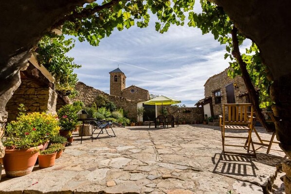 A courtyard with stone flooring, potted plants, outdoor furniture, and a view of a historic building with a tower.