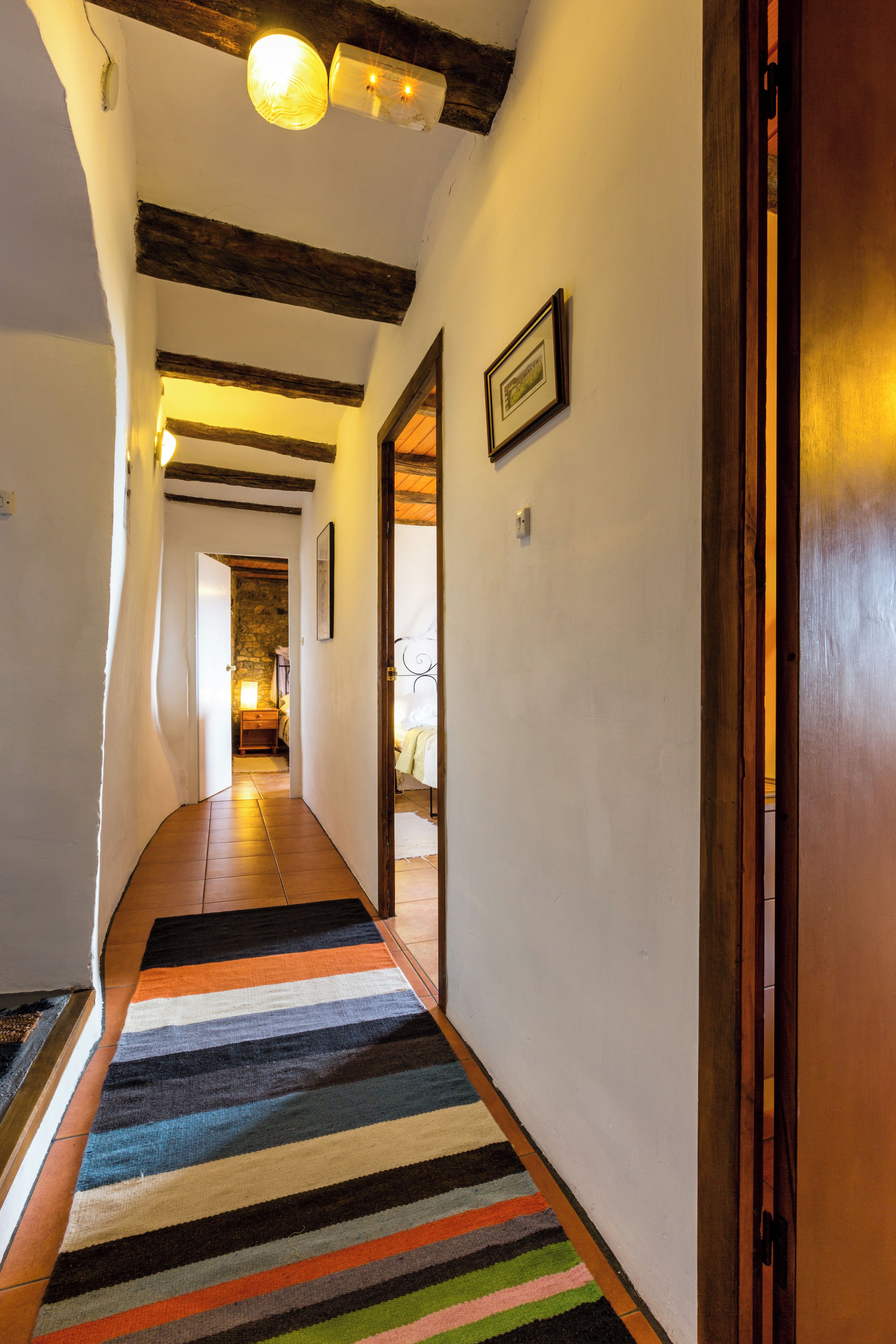 Un pasillo con alfombra a rayas, vigas de madera, un cuadro enmarcado y una puerta que da a otra habitación.