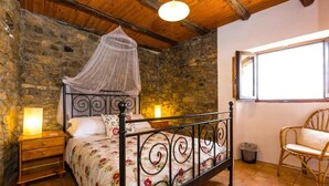 Quarto com parede de pedra, cama com estrutura de metal, cadeira de vime e janela.