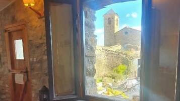 Habitació amb paret de pedra, finestra amb vista a un edifici i una cadira amb coixins.