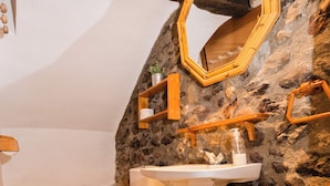Casa de banho rústica com parede de pedra, vigas de madeira, pia branca, sanita e espelho.