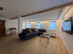 Una sala de estar moderna con un sofá oscuro, una mesa de centro de vidrio, piso de madera y un travesaño de madera en el techo.