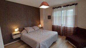 A bedroom with a bed, bedside lamps, a dark-colored armchair, and a window with curtains. - Holiday Home 'Gîte Du Mansois' with Private Terrace, Private Garden and Balcony (Clairvaux-d'Aveyron)