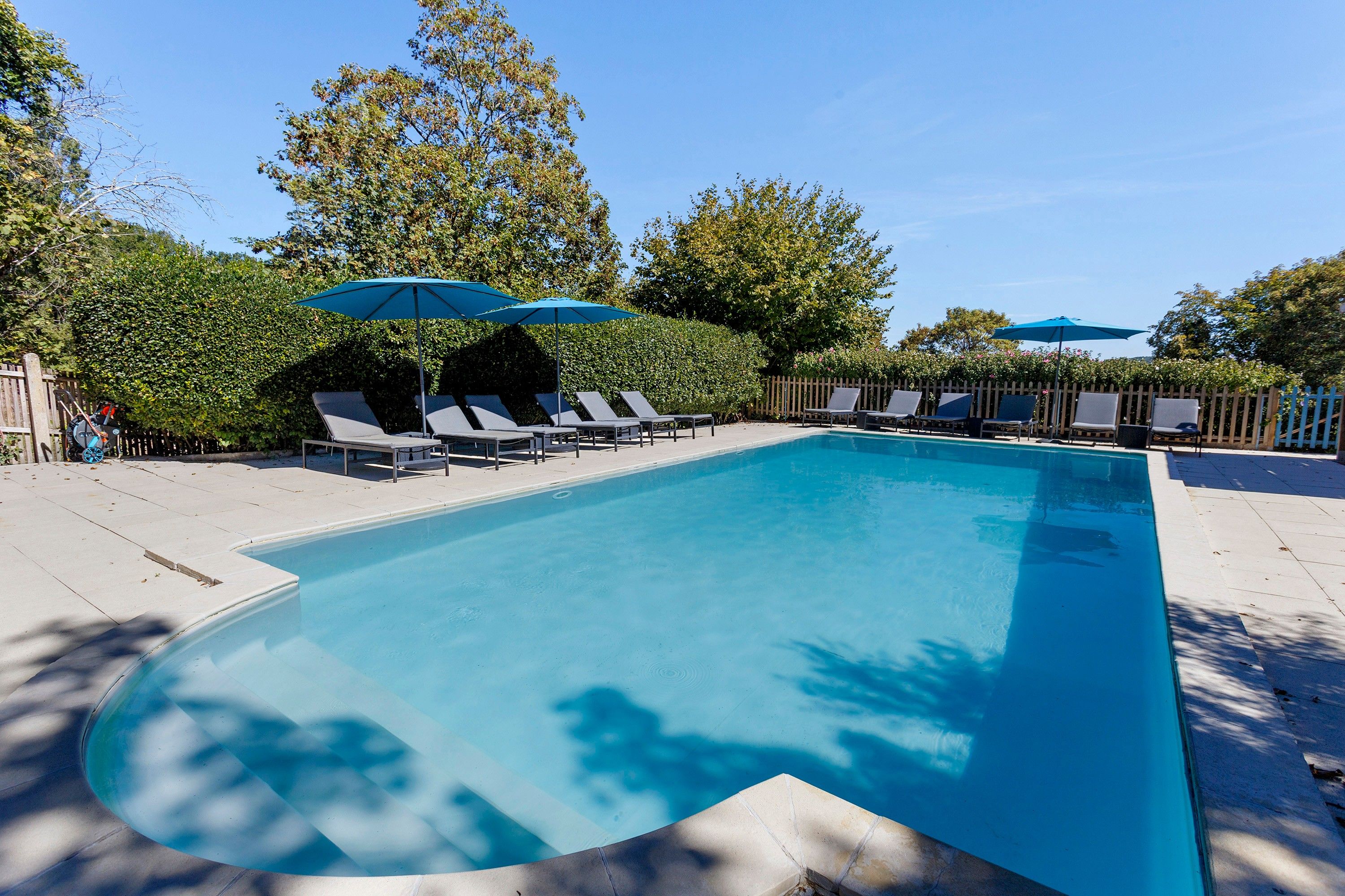 Ein rechteckiger Swimmingpool mit einer gefliesten Kante, umgeben von Liegestühlen und Sonnenschirmen, mit einem Holzzaun im Hintergrund.