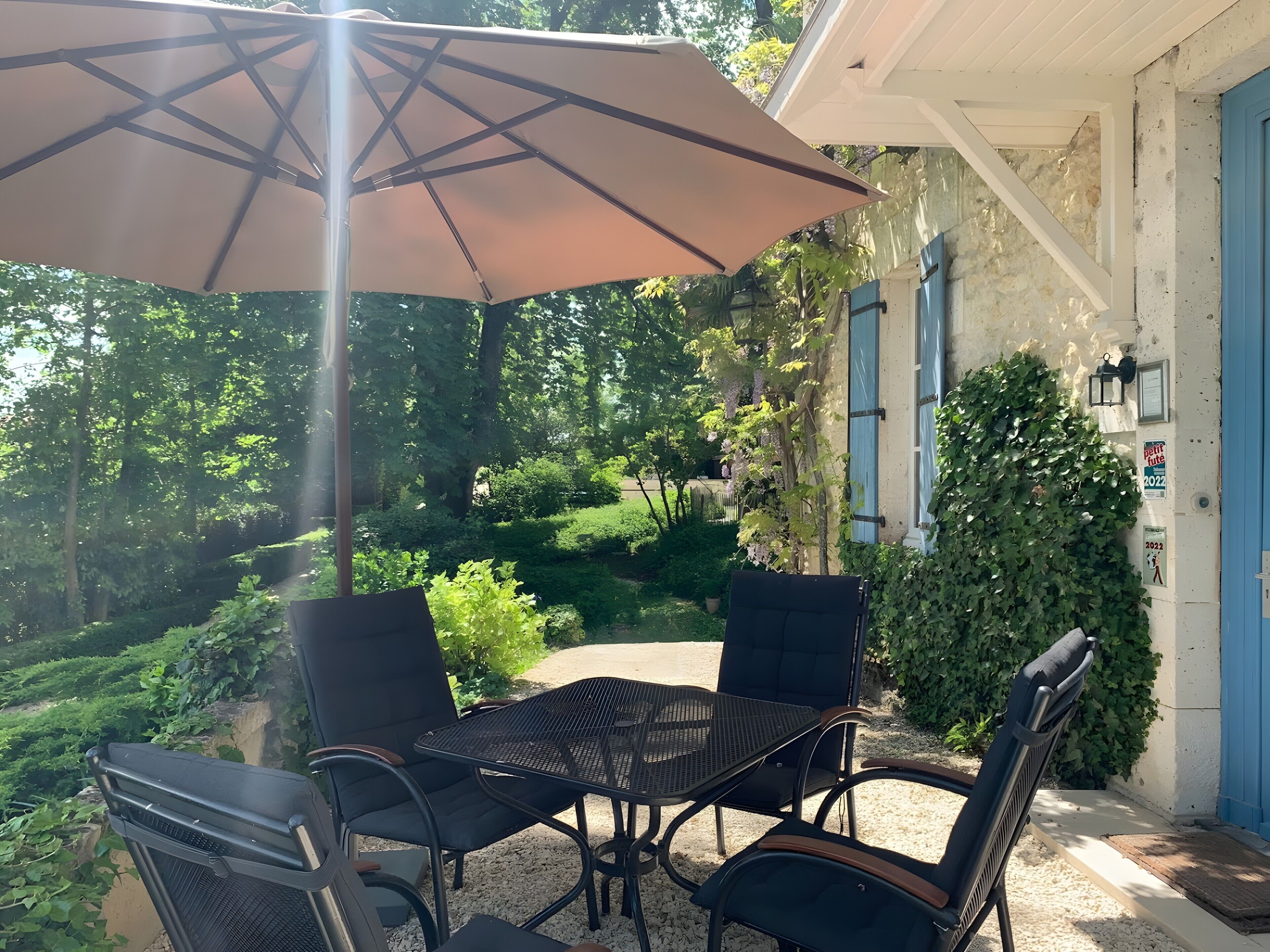 Eine Terrassengruppe im Freien mit Tisch, Stühlen und einem großen Sonnenschirm, mit Blick auf einen Garten mit Bäumen und einer Hecke.