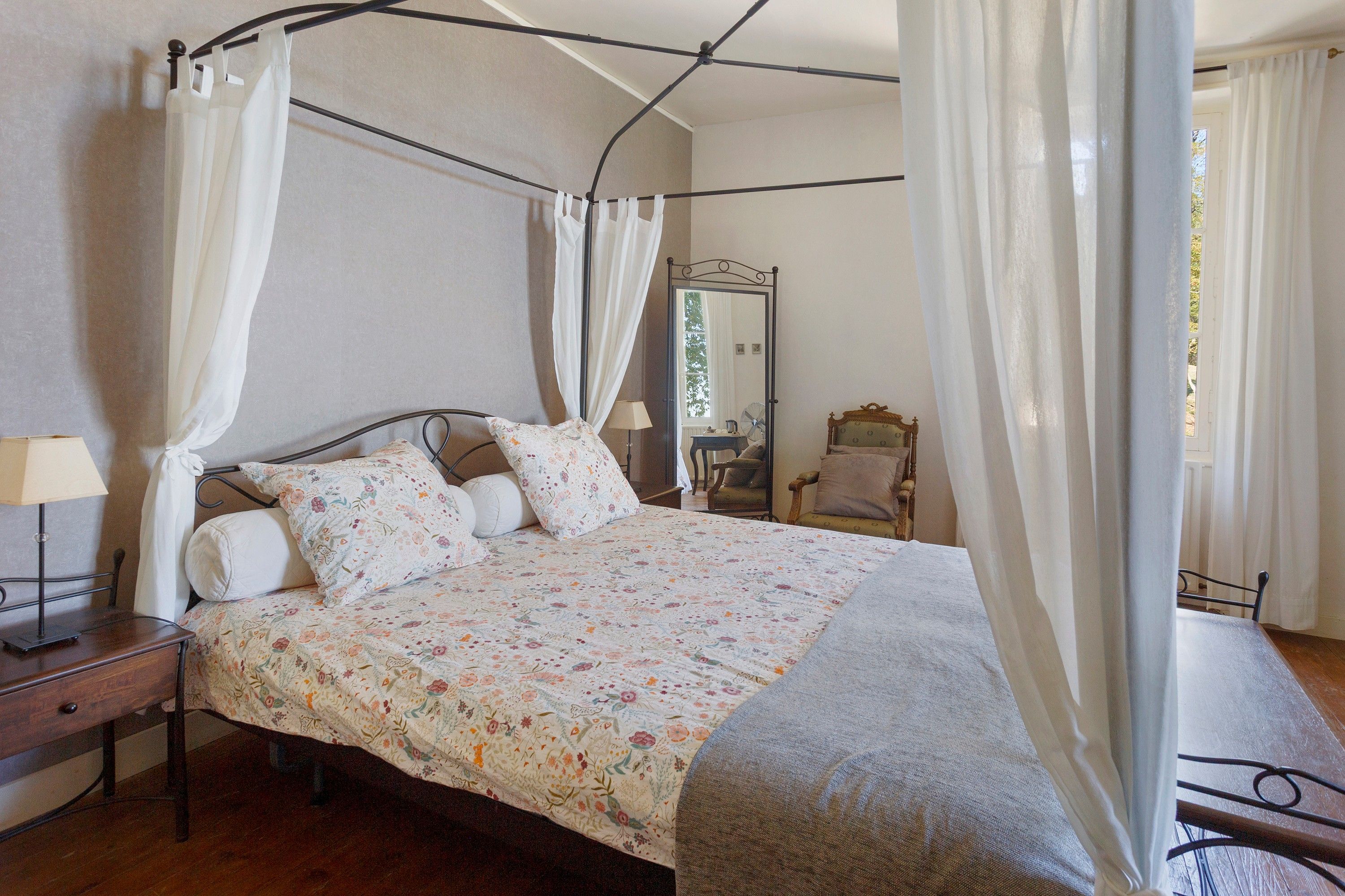 Un lit à baldaquin avec une couvre-lit à motifs floraux, une table de chevet avec une lampe et une chaise dans une pièce dotée d’un grand miroir et de rideaux.
