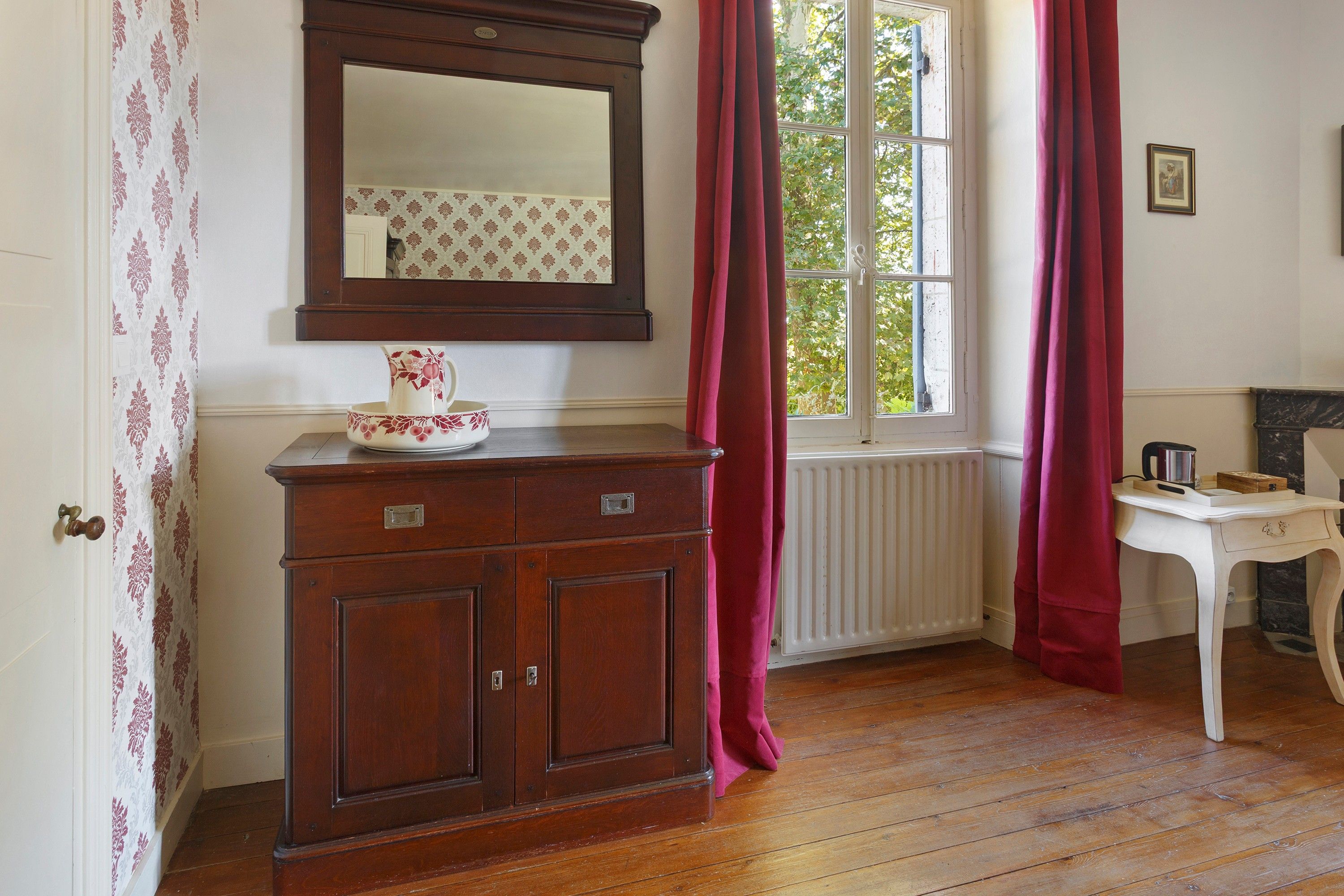A room with a wooden cabinet, a mirror, a window with curtains, a radiator, and a small table with a kettle.