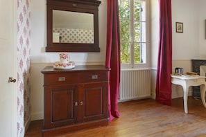 Una habitación con un armario de madera, un espejo, una ventana con cortinas, un radiador y una pequeña mesa con una tetera.