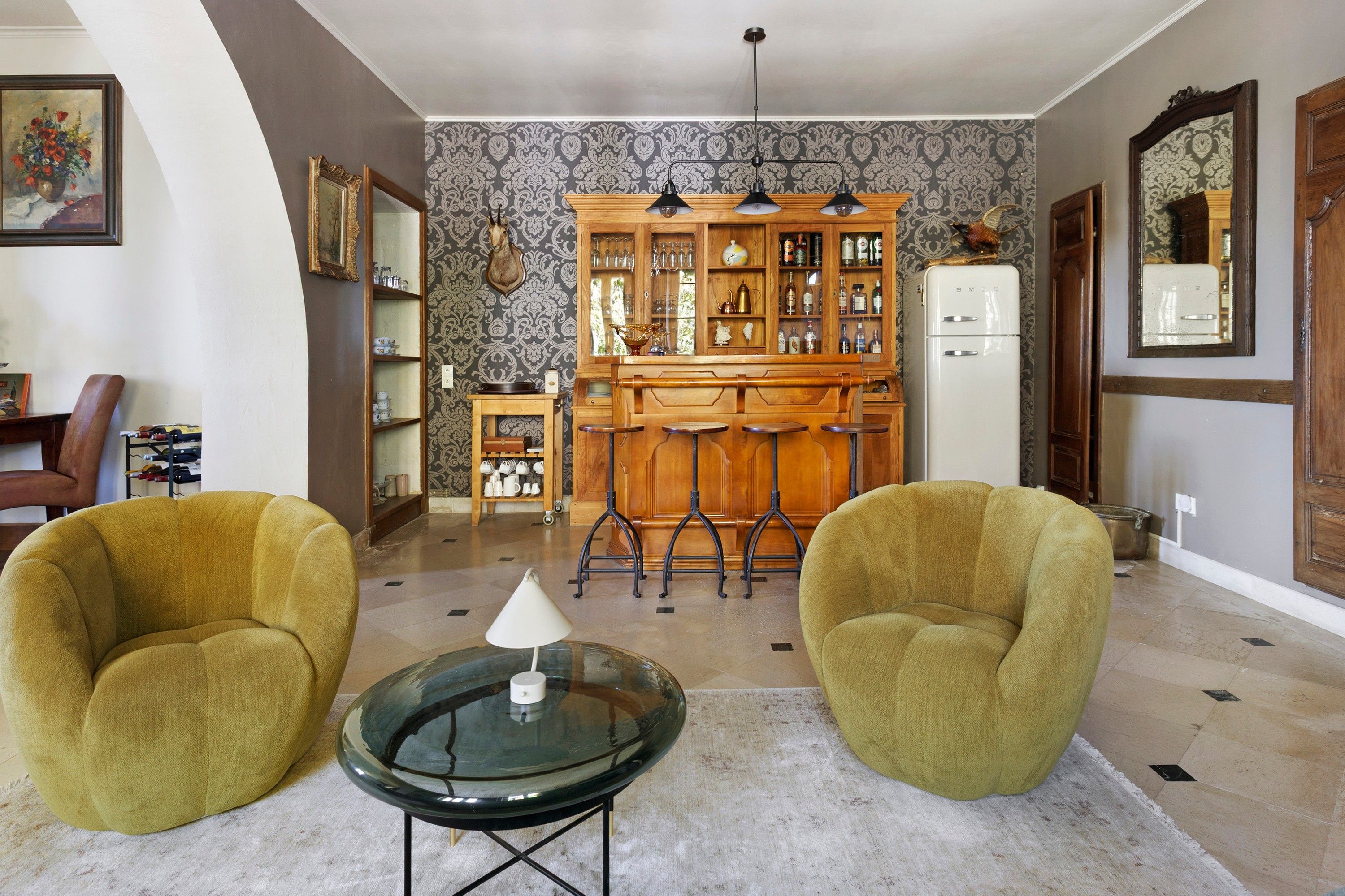 Una sala de estar con dos sillones, una mesa de centro de vidrio, un mueble de madera con puertas de cristal y un refrigerador blanco.